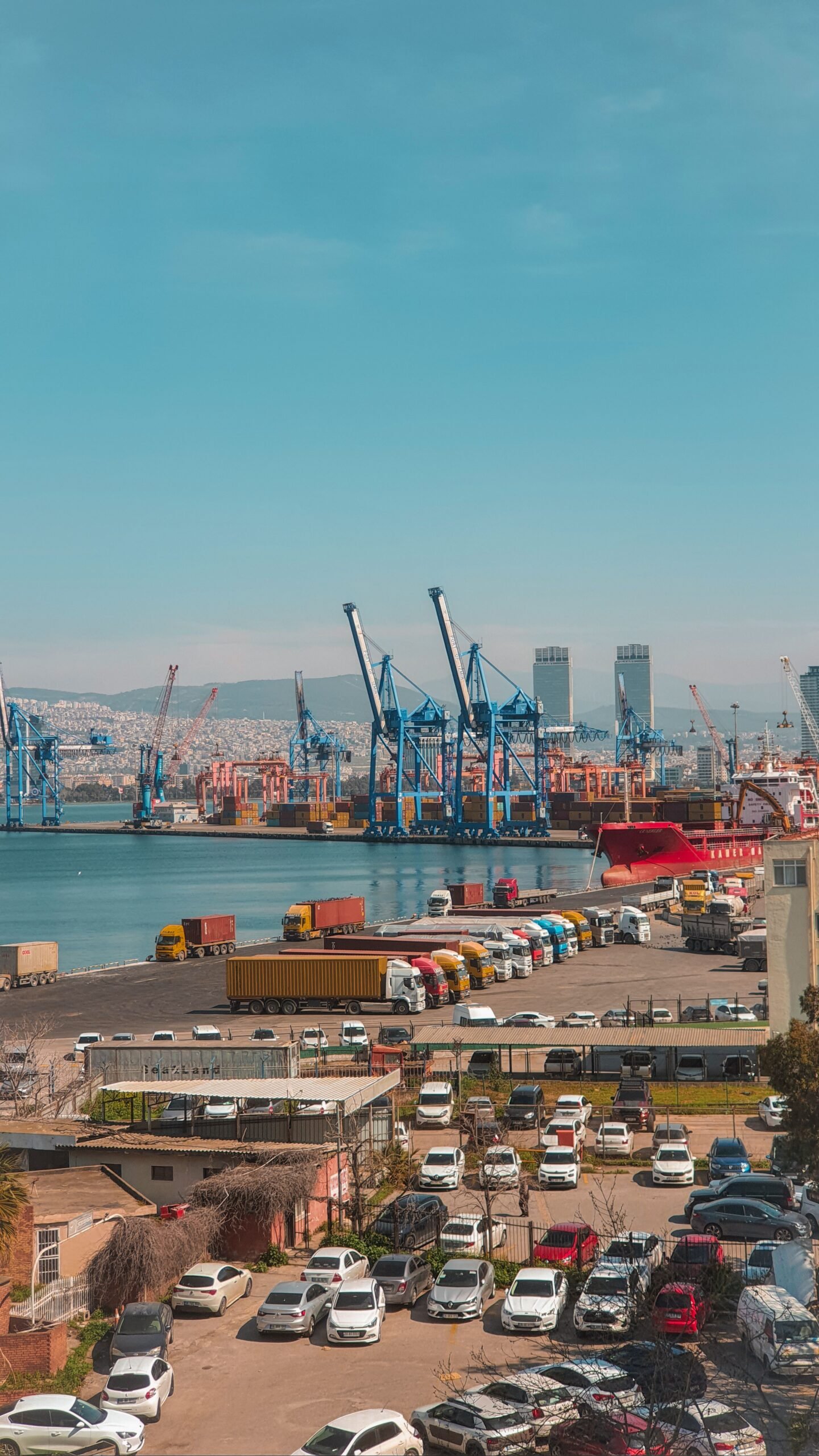 About A busy day at the İzmir harbor showcasing cargo ships, cranes, and vehicles in an urban setting.
