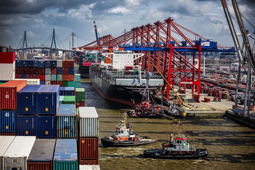 Vibrant port scene with a large cargo ship being guided by tugboats, flanked by towering red cranes and stacks of multicolored shipping containers under a dynamic sky.